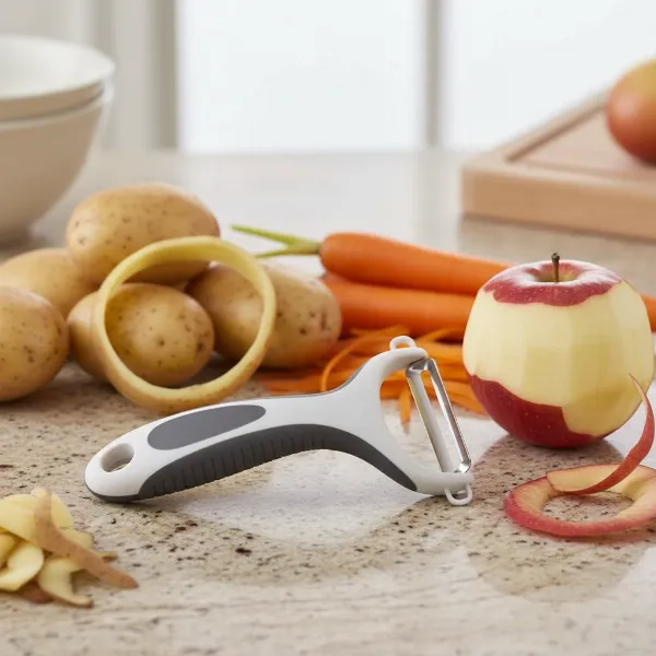 A Zyliss Smooth Glide Swivel Peeler alongside various fruits and vegetables like potatoes, carrots, and apples, illustrating its versatility and European design.